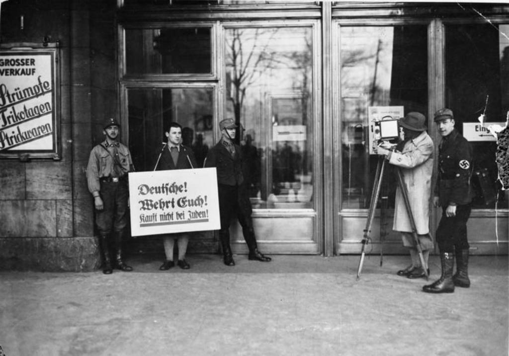 Vor einem jüdischen Geschäft stehen Menschen mit einem Schild mit der Aufschrift 