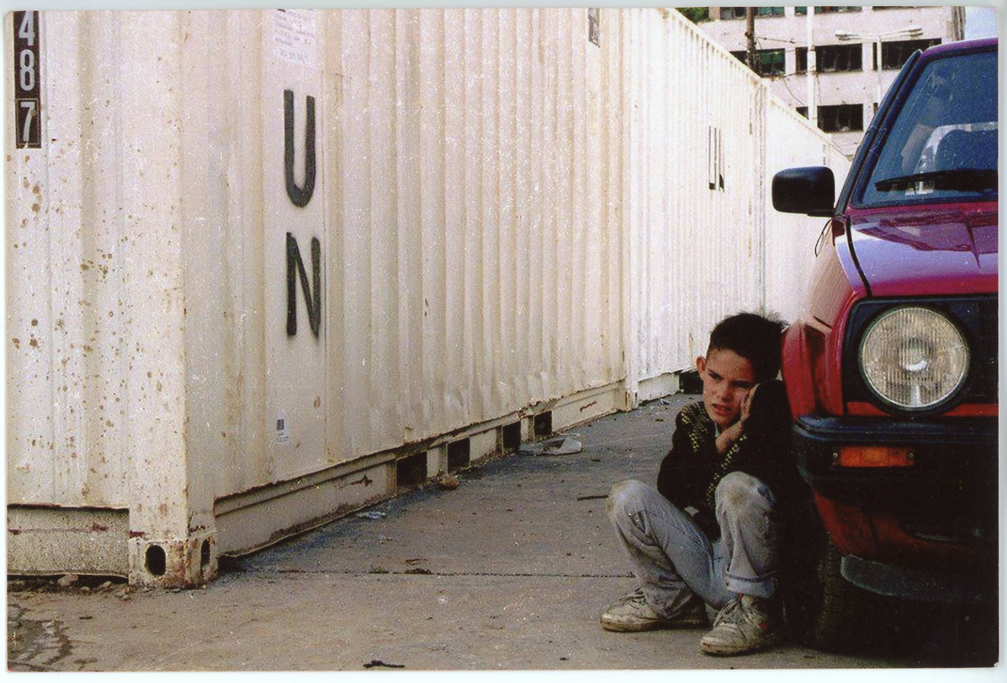 Kind versteckt sich hinter einem Auto. Im Hintergrund ist ein weißer UN Container zu sehen.